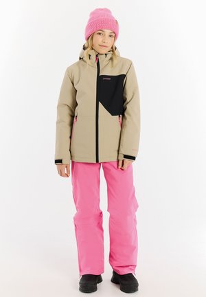 Hellbeige, wasserdichte Jacke mit schwarzen Akzenten, kombiniert mit leuchtend rosa Schneehosen und einer rosa gestrickten Mütze. Schwarze Stiefel runden den Look ab.