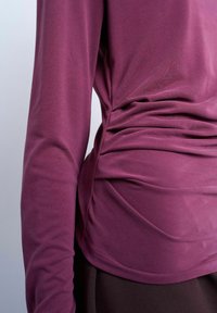 Haut prune ajusté à manches longues avec détails froncés sur les côtés, porté par une personne sur un fond clair uni.