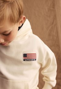 Krämsfärgad hoodie med en framficka och ett broderat märke med en amerikansk flagga och "SUPERDRY"-logotyp i blått och rött. Mjuk textur.