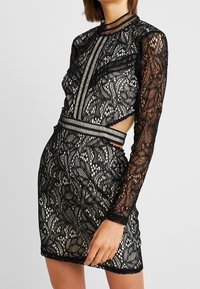 Black lace long-sleeve dress featuring a fitted design with floral patterns, underlay in cream, and cut-out side accents with striped trim.