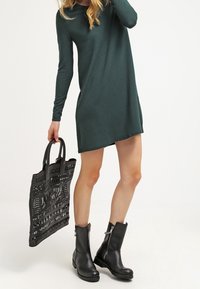 Czarna sukienka z długim rękawem w ciemnozielonym kolorze, sięgająca powyżej kolan; w połączeniu z czarnymi botkami za kostkę z zamkiem błyskawicznym oraz czarną torebką z fakturowanej tkaniny.