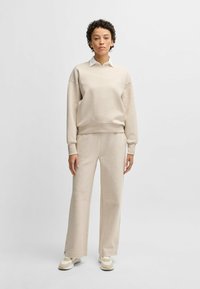 Felpa beige con polsini e orlo a coste, indossata sopra una camicia bianca con colletto; pantaloni della tuta beige abbinati con dettaglio a pressione all'orlo.