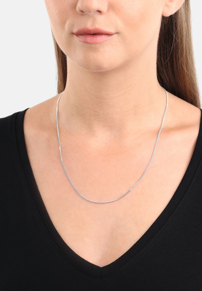 Silverhalsband med en slät, flexibel design, som har en fin kedjetextur som hänger precis ovanför en svart t-shirtkrage.