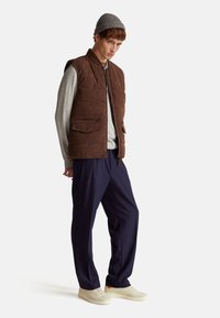 Hnědá kordová vesta s vysokým límcem, dvěma předními kapsami, pod ní šedá košile s dlouhým rukávem, námořnické kalhoty a béžové tenisky.