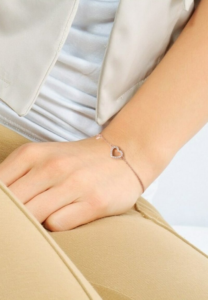 Rose gold bracelet with a heart-shaped charm, adorned with clear gemstones, positioned on a wrist against a neutral background.