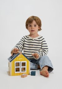 Casa di legno giocattolo con strisce gialle e tetto grigio. Bambino in maglione a righe interagisce con essa su uno sfondo bianco.