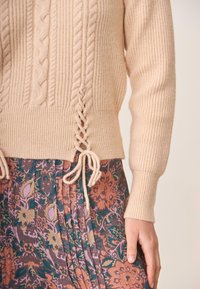 Sweater beige de punto trenzado con textura acanalada y lazos de flecos en el dobladillo, combinado con una falda plisada de estampado floral en tonos terrenales.