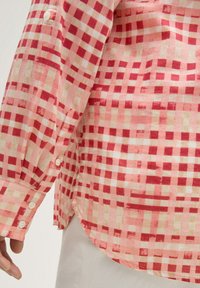 Chemise à carreaux rouge et rose clair en tissu léger, avec un poignet boutonné et un ourlet courbé, offrant une texture lisse.