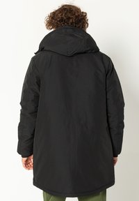 Zwarte gewatteerde jas met een capuchon, met een gladde textuur en een lange snit, gezien vanaf de achterkant. Minimalistisch ontwerp zonder zichtbare patronen.