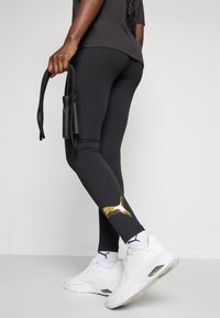Fekete, fitted dizájnnal rendelkező leggings, arany logó részletekkel a bokán. Fehér tornacipővel viselve, és ugrókötél van a kezében.