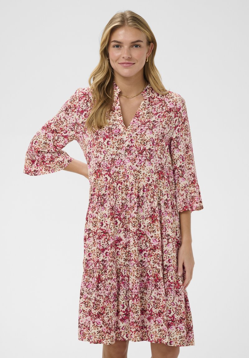 Jeune femme portant une robe florale rose, rouge et beige, longueur genou, à manches trois-quarts, debout avec une main sur la hanche.