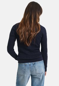 Marineblauer Kabelstrickpullover mit geripptem Saum und Manschetten, mit taillierter Silhouette und langen Ärmeln, kombiniert mit hellblauen Jeans.