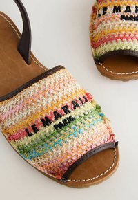 Sandales à bouts ouverts avec des brides tissées multicolores, un bord en cuir marron et un texte noir brodé sur la partie avant supérieure.