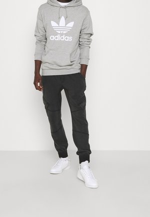 Mężczyzna w szarej bluzie z kapturem Adidas, czarnych spodniach typu cargo i białych sneakersach Adidas stoi na tle prostego jasnego tła.