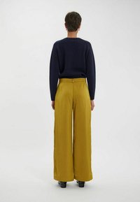 Un pull en maille bleu marine et un pantalon à taille haute, large, en velours côtelé moutarde, portés avec des bottes noires à talons, vus de dos.