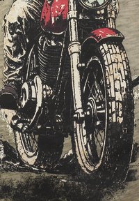 Primo piano della ruota anteriore e del motore di una motocicletta con un guidatore che indossa pantaloni beige, parafango rosso e stivali neri su una superficie texturizzata.