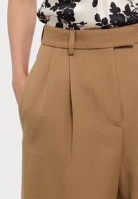 Beige tailored trousers with a textured finish, front pleats, side pockets, and a waistband with a belt loop. Fabric has a subtle sheen.