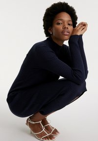 Navy blauer, gerippter Strickkleid mit hohem Kragen und langen Ärmeln, ausgestattet mit seitlichen Schlitzen. Kombiniert mit weißen, schnittigen High-Heels.