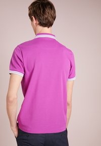 Homme portant un polo à manches courtes rose vif avec un col et des poignets à rayures blanches, de dos, les mains dans les poches de son pantalon sombre.