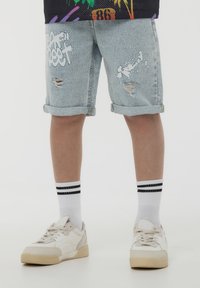 Bermuda shorts in denim azzurro chiaro con dettagli strappati, orlo arrotolato, abbinati a calze bianche con strisce nere e sneakers bianche.
