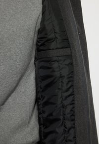 Tela de sudadera gris por fuera, forrada con un acolchado negro suave. Presenta un bolsillo lateral y un detalle de cremallera a lo largo del borde.