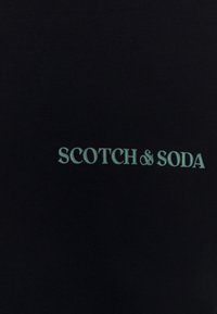 Camiseta de tela negra con textura acanalada, que presenta el bordado "SCOTCH & SODA" en un tono azul claro. Diseño clásico de cuello redondo.