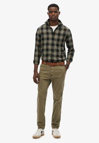 Geruite button-up shirt in donkergroen en geel, gecombineerd met kaki broeken en witte sneakers met grijze accenten. Bruine riem met een gestructureerde afwerking.