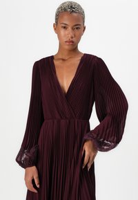 Burgundy pleatattu mekko, jossa on syvä V-pääntie, pitkät hihat ja läpinäkyvät pitsikoristeet hihansuissa. Nesteinen, teksturoitu kangas parantaa liikettä.