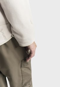 Helle beige Fleecejacke mit glatter Textur und einem Logo-Tag, kombiniert mit olivgrünen Hosen, die eine Seitentasche und eine weiche Haptik aufweisen.