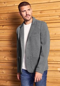 Harmaat vaatteet sisältävät rakenteellisesti muotoillun designin, jossa on viilletty kaulus ja kaksi etutaskua, valkoisen paidan ja sinisten farkkujen päällä.