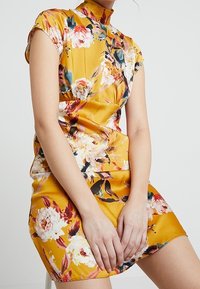 Robe florale en satin jaune avec un col montant et des manches courtes. Présente de grands motifs floraux colorés et une silhouette ajustée.
