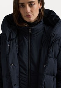 Zelena puffer jakna s visokim ovratnikom i velikim, strukturiranim kapuljačom. Ima prednji zatvarač i dugmad s bočne strane. Glatka tekstura.