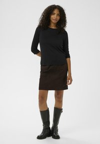 Haut noir à manches longues, mini-jupe en suède marron avec poches avant, et bottes noires en cuir jusqu'aux genoux. Design simple et ajusté.