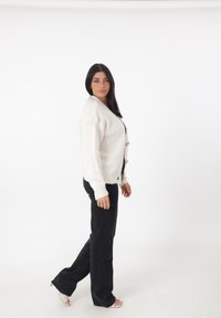 Witte gebreide cardigan met een V-hals, voorzien van houten knopen en ribbelmanchetten, gecombineerd met zwarte flared broeken en sandalen met doorzichtige hakken.