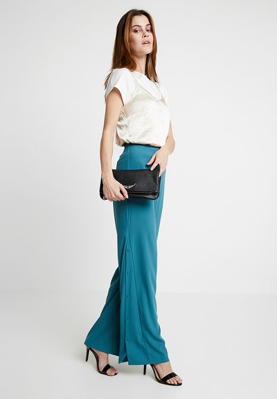 Pantaloni a gamba larga color teal con dettagli a bottoni lungo il lato, abbinati a un top in satin chiaro e una pochette nera. Il modello indossa sandali neri con tacco.