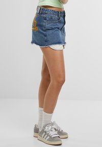 Denim mini skirt with a frayed hem, blue color, featuring an embroidered yellow logo on the back pocket. Paired with gray sneakers and white socks.