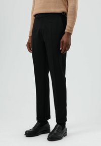 Zwarte, op maat gemaakte broek met een rechte pijp, een gladde textuur, riemlussen en omslagen, gecombineerd met zwarte loafers.