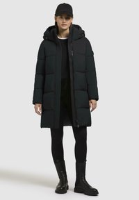 Zwarte pufferjas met een capuchon, uitgevoerd met een gestikte design, rits aan de voorkant, zijzakken, gedragen over een zwarte top en leggings, gecombineerd met zwarte laarzen.