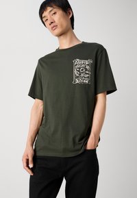 Olivenfarvet bomulds t-shirt med korte ærmer, prydet med et hvidt grafisk design på venstre bryst. Blød tekstur og afslappet pasform.