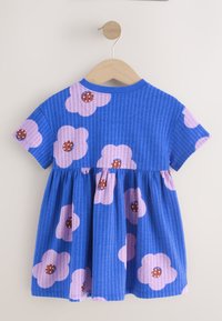 Vestido corto azul para bebé con dibujos de flores moradas, mangas cortas y cintura fruncida, colgado en una percha de madera contra una pared blanca.