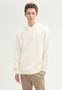 Kremowy hoodie z kangurzą kieszenią, kapturem z troczkami i subtelnie wytłoczonym logo. Wykonany z miękkiej tkaniny o gładkiej teksturze.