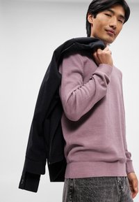 Lyse lilla sweater med en tekstureret finish, parret med en sort jakke draperet over skulderen. Modellen bærer grå jeans.