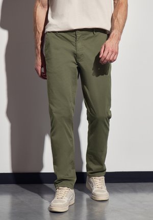 Green cotton chinos with a straight cut, flat front, and side pockets, paired with beige sneakers on a concrete floor.
