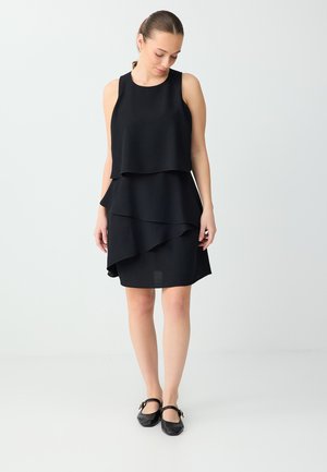 Młoda kobieta o jasnej cerze i brązowych włosach związanych w kucyk, ubrana w warstwową czarną sukienkę bez rękawów i czarne baleriny Mary Jane, stojąca na białym tle.