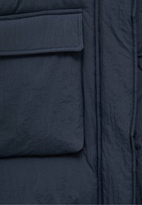 Gros plan d'une poche de manteau en tissu froissé bleu foncé avec un rabat et un détail de couture verticale.