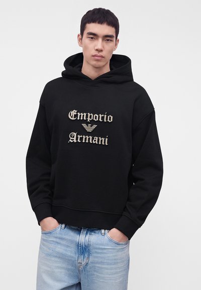 Νεαρός άνδρας με κοντά μαύρα μαλλιά, που φοράει μαύρο φούτερ Emporio Armani και ανοιχτό μπλε τζιν, χέρια στις τσέπες, ουδέτερο φόντο.