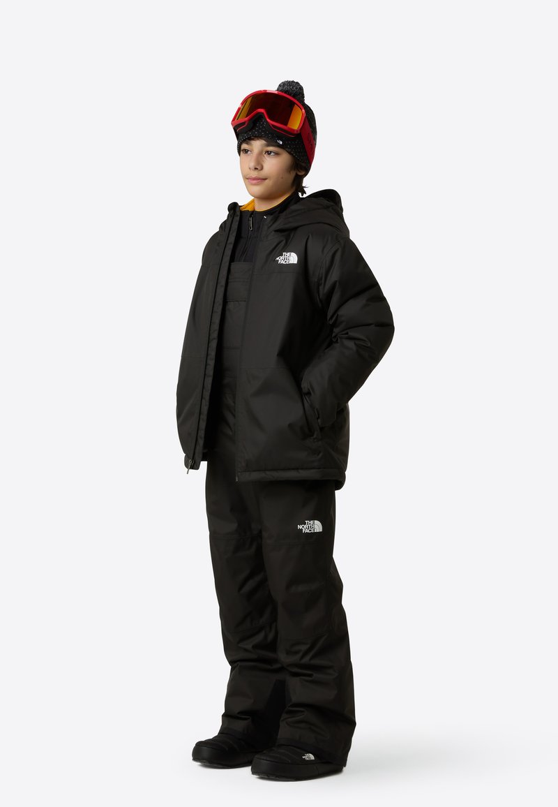 Svart isolerad snöjacka och överdragsbyxor med The North Face-logotyp. Barnet har på sig en stickad mössa och stora skidglasögon. Texturerad tygfinish.
