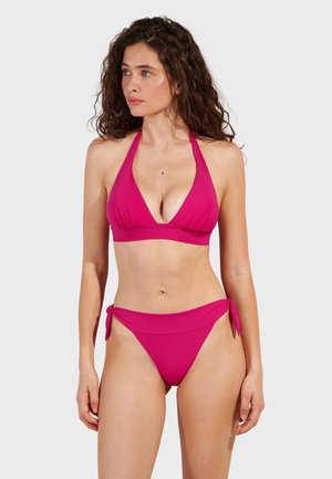 Žena s kovrčavom kosom u dvočlanom magenta bikiniju s halter topom i donjim dijelom s bočnim vezicama, gleda lijevo na bijeloj pozadini.