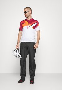 Maillot de cyclisme à manches courtes et zippé en blanc avec des motifs diagonaux violets, orange et noirs ; associé à un pantalon noir et un casque blanc.