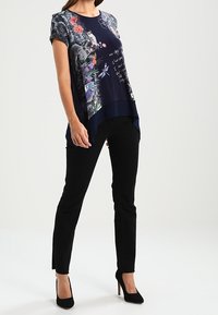 Kvinna iklädd en marinblå blommig grafisk t-shirt, svarta slim fit-byxor och svarta högklackade skor, stående mot en enhetlig bakgrund.